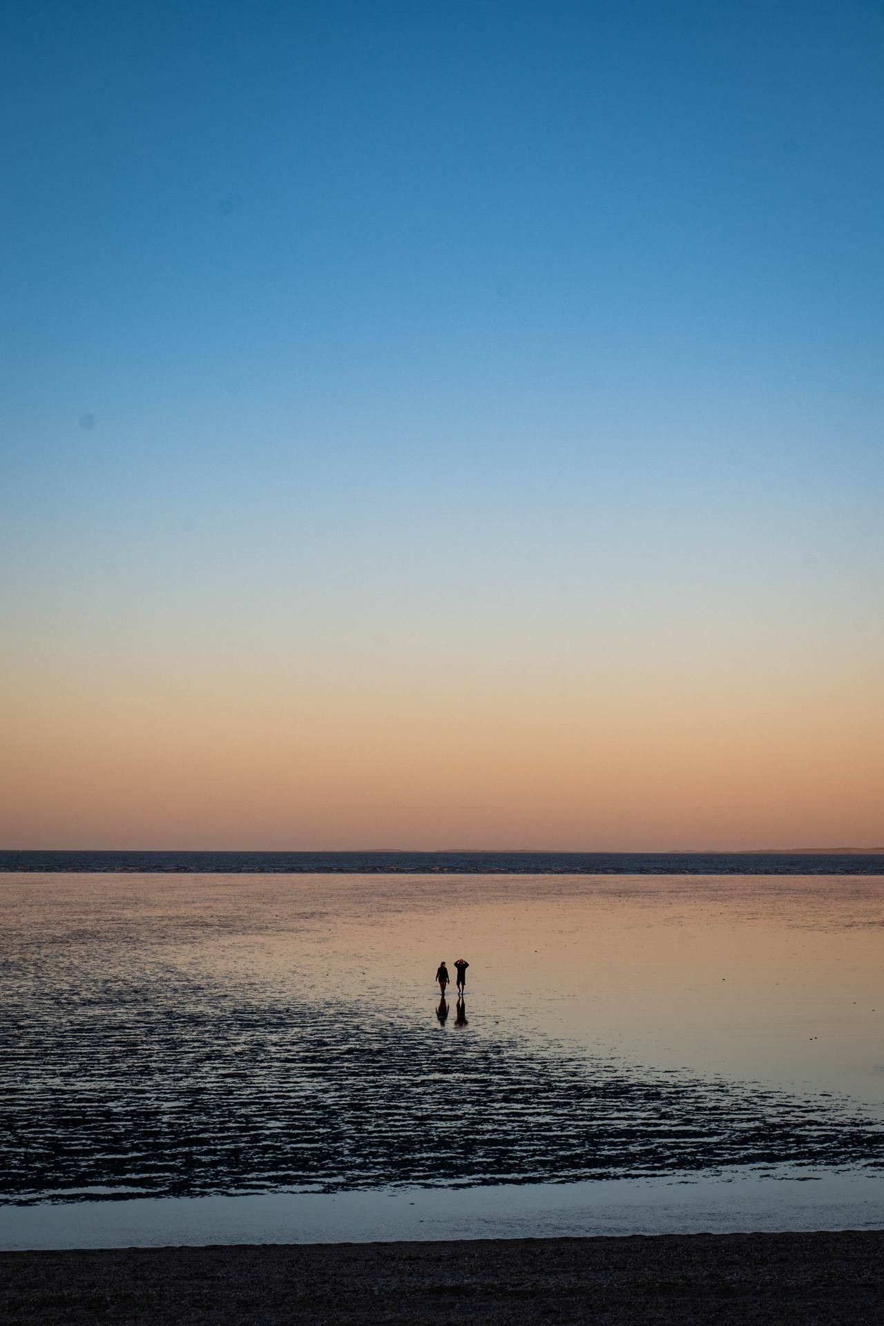 Two people walking across shallow tidal water beneath a pastel evening sky