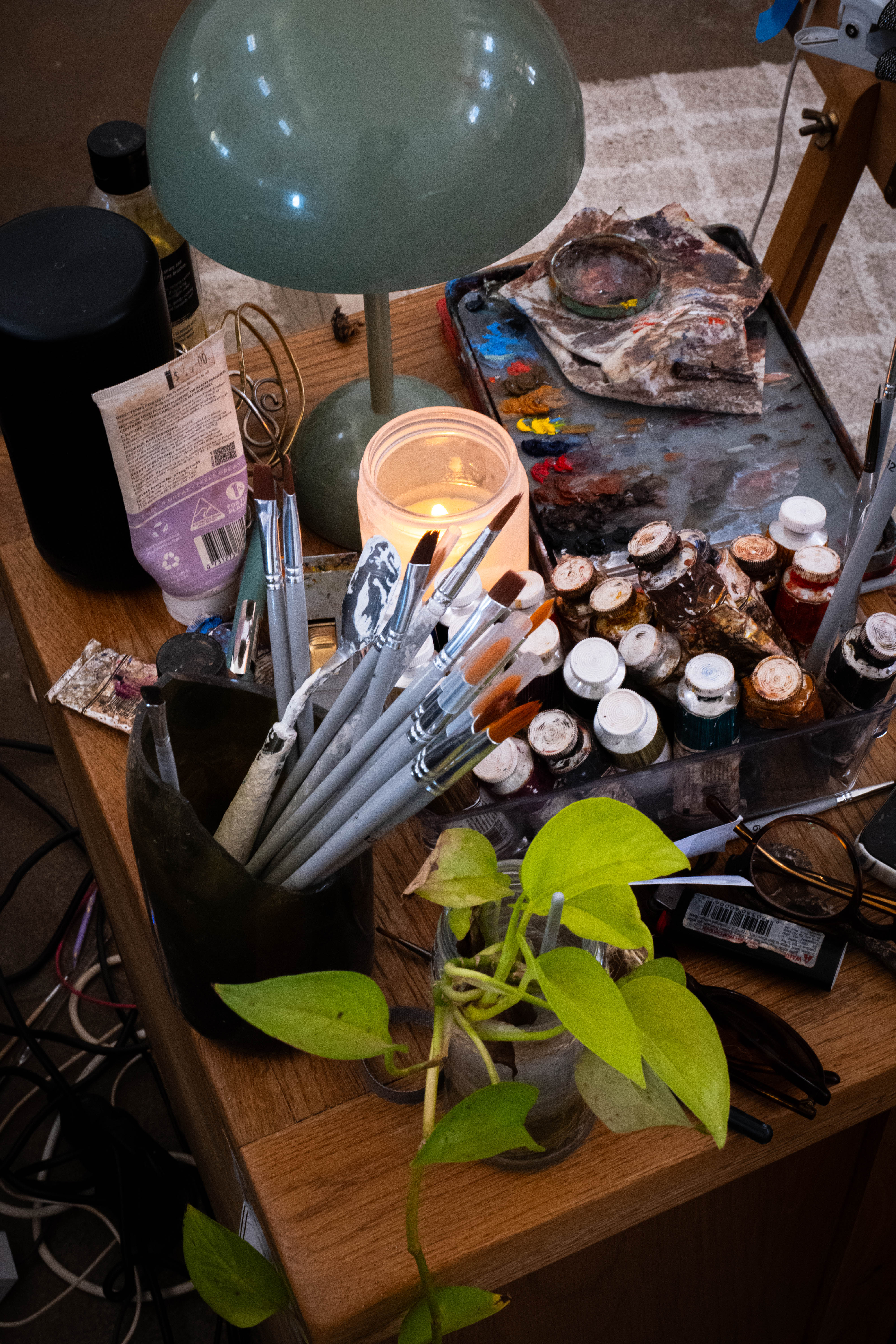 An artist’s table filled with brushes, paints, a lamp and a trailing plant