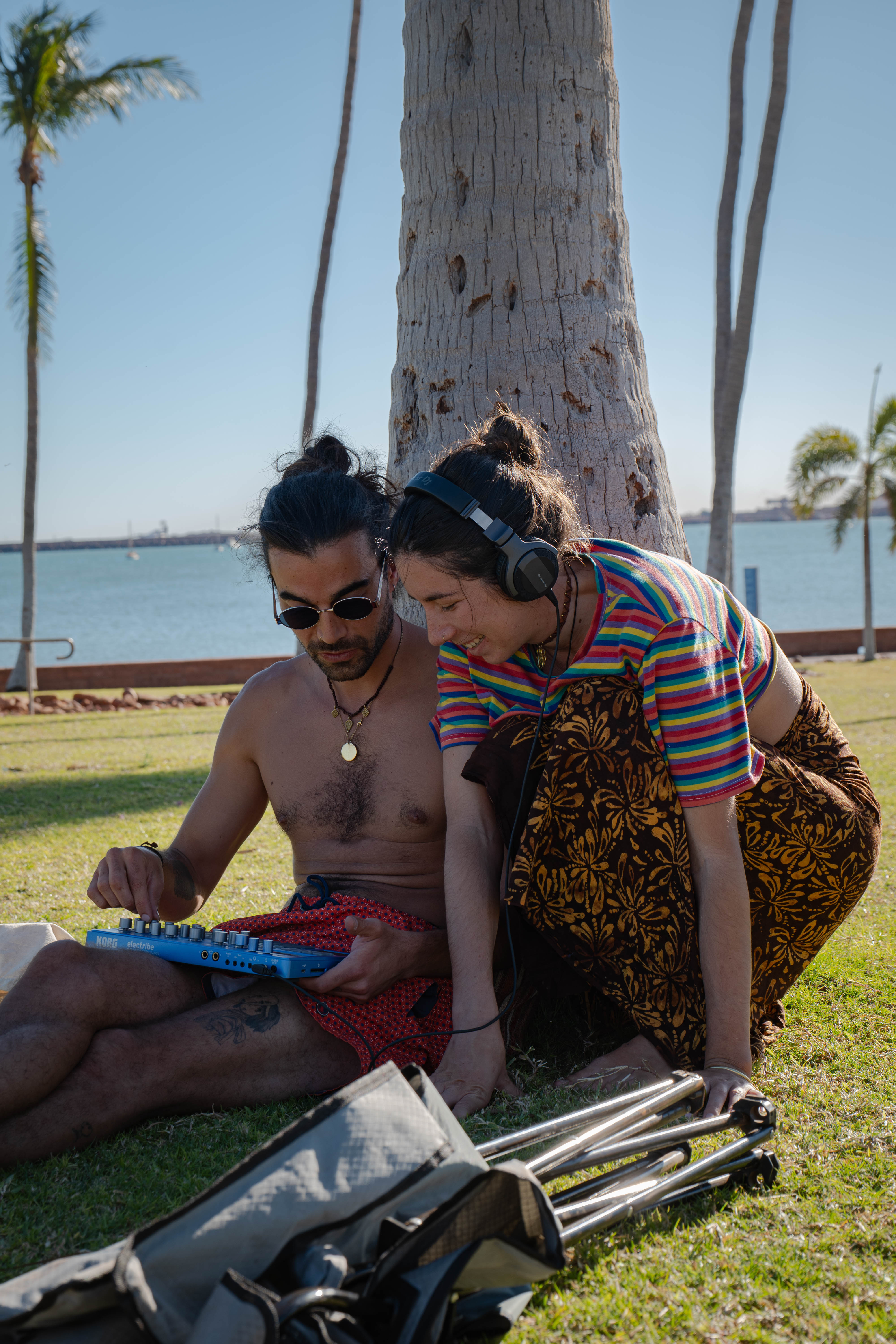 Two musicians adjusting a small synthesizer together on waterfront grass
